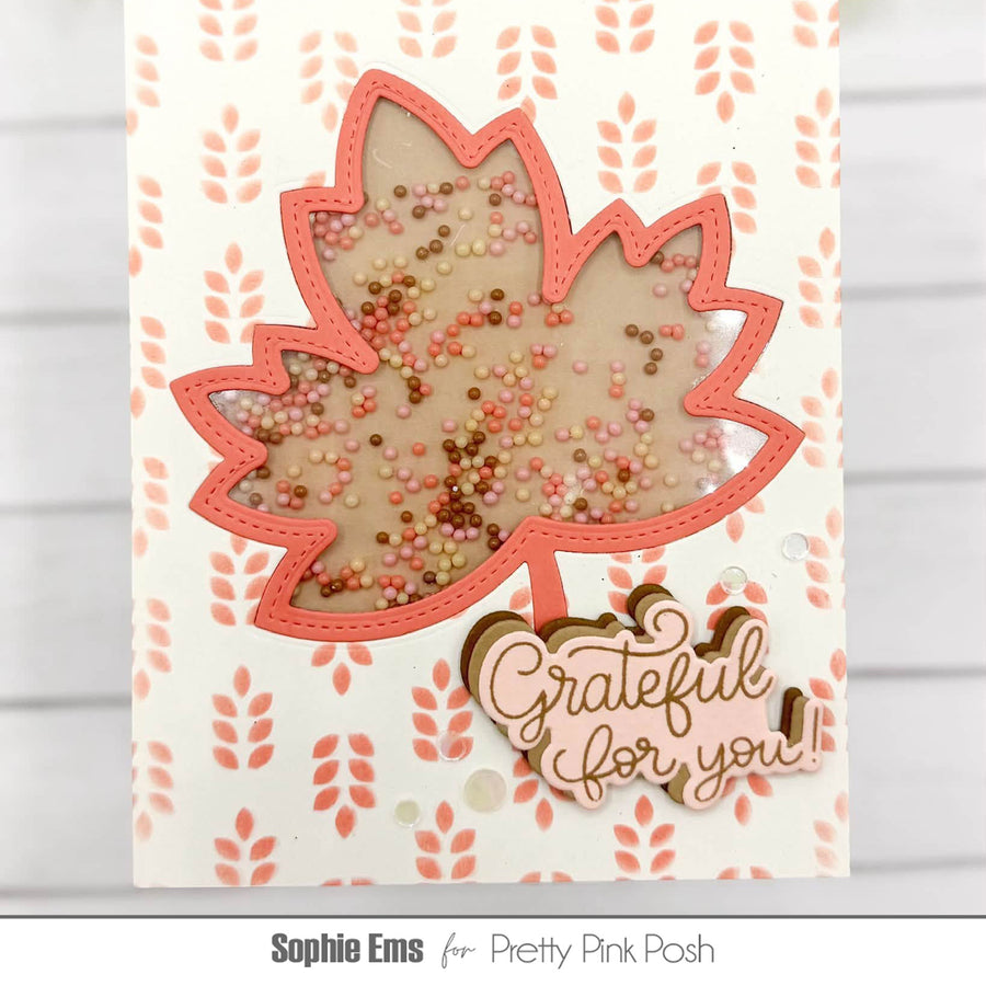 A handmade greeting card featuring a large maple leaf with a transparent center filled with multicolored sequins, surrounded by a coral-pink stitched border. Below the leaf, the words 'Grateful for you!' are written in metallic gold script.