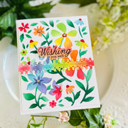 A handmade greeting card with colorful watercolor-style flowers and green leaves, featuring the phrase 'Wishing you well' in script. A rainbow-striped decorative band with small pom-poms runs horizontally across the center.