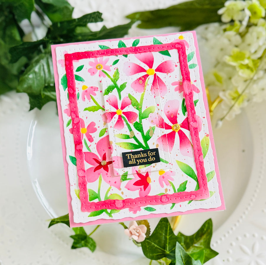 A handmade floral greeting card with pink and red flowers, green leaves, and a small black card reading 'Thanks for all you do' in yellow text, styled on a white plate with green leaves and pale flowers in the background.