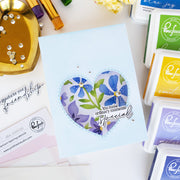 A handmade light blue greeting card featuring a heart-shaped floral stamp design with blue and purple flowers, green leaves, and the text 'You make ordinary moments special.' The card is styled on a white surface with crafting supplies, including multiple Painful Punch ink pads, a gold dish with rhinestones, and paintbrushes.