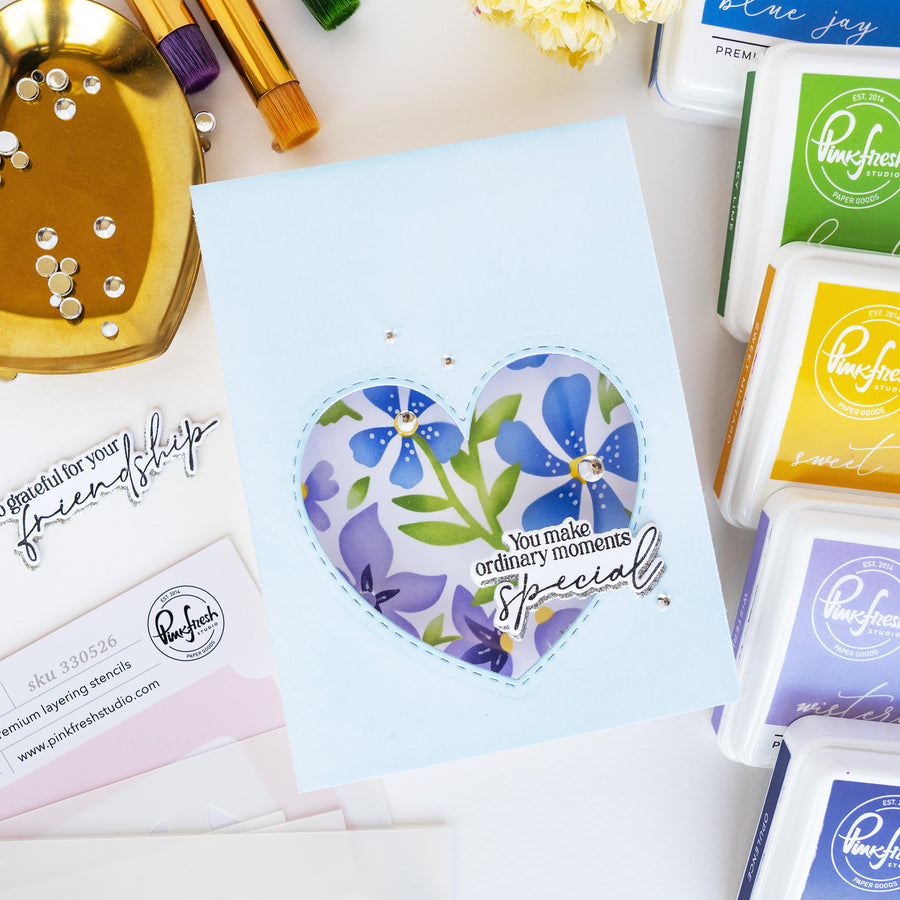A handmade light blue greeting card featuring a heart-shaped floral stamp design with blue and purple flowers, green leaves, and the text 'You make ordinary moments special.' The card is styled on a white surface with crafting supplies, including multiple Painful Punch ink pads, a gold dish with rhinestones, and paintbrushes.