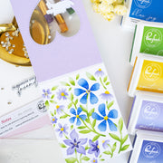 A flat-lay of craft supplies featuring a floral stamp card with blue, purple, and green flowers, alongside Ranger ink pads in blue, yellow, and purple, and a gold dish with small white flowers on a white surface.