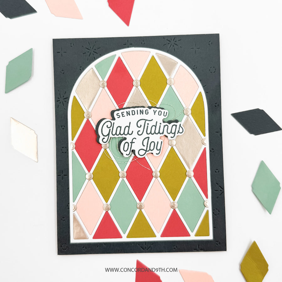 A handmade layered card featuring a black embossed border, a white arched background, and a colorful diamond pattern in red, green, pink, and gold. The center has a die-cut layer reading 'Sending You Glad Tidings of Joy' with a small bow.