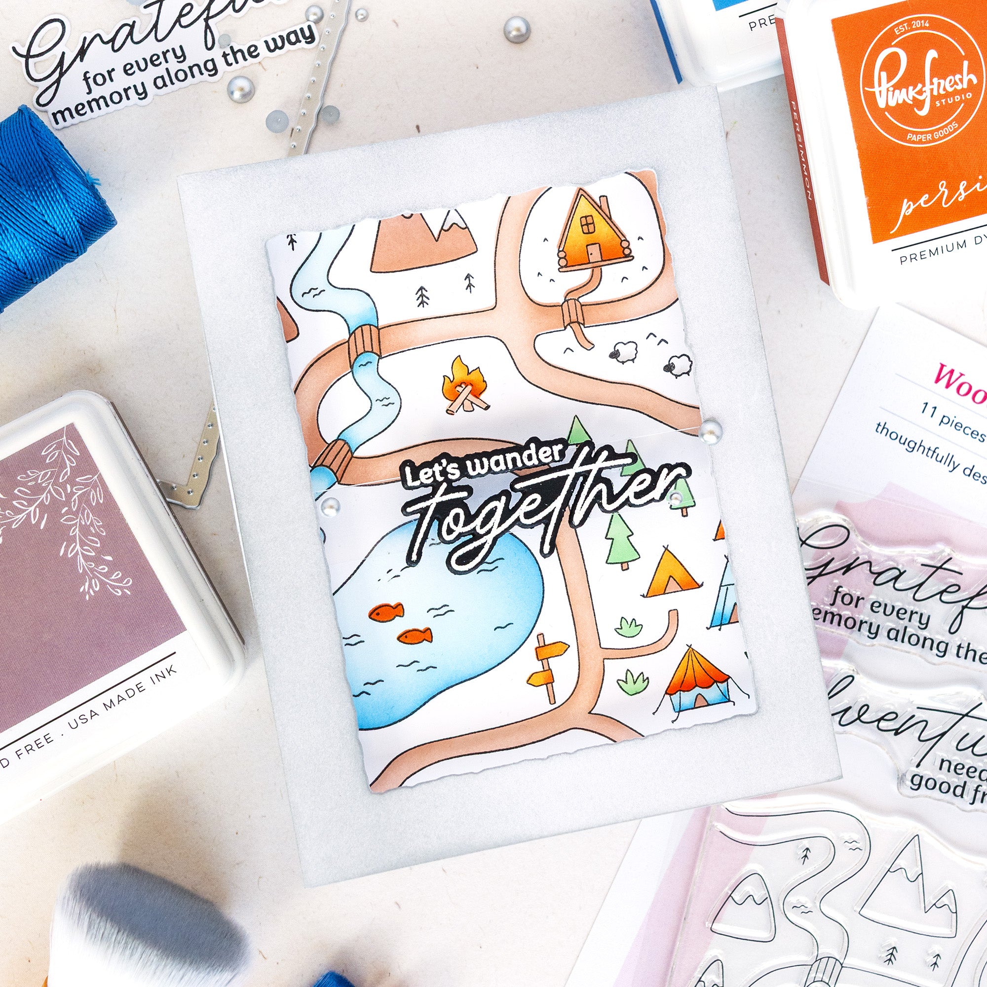 A flat-lay of a handmade greeting card featuring a whimsical illustrated map with the phrase 'Let's wander Together' in stylized lettering. The card is surrounded by crafting supplies, including an orange Pine Tech premium ink pad, a blue ink pad, and other card-making materials on a white surface.