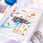 A close-up of a handmade greeting card featuring a whimsical illustrated map with a blue pond, orange trees, and the phrase 'Let's wander together' in raised black lettering. The card is layered on a light gray cardstock base, with other crafting materials partially visible around it.