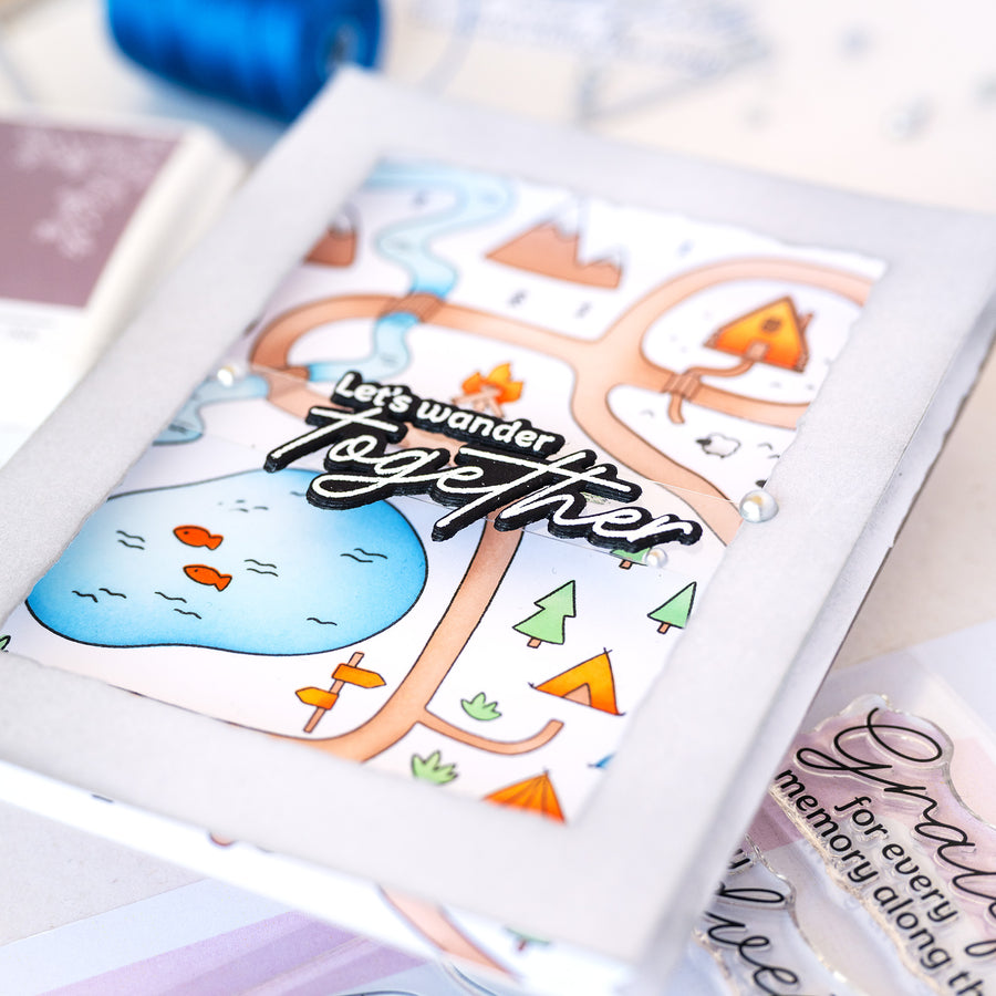 A close-up of a handmade greeting card featuring a whimsical illustrated map with a blue pond, orange trees, and the phrase 'Let's wander together' in raised black lettering. The card is layered on a light gray cardstock base, with other crafting materials partially visible around it.