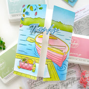 A close-up of a handmade greeting card featuring a colorful illustration of a pink rowboat on blue water, with the word 'Thanks' in large script across the front. The card is layered over other craft supplies, including pink and green ink pads and clear stamps.