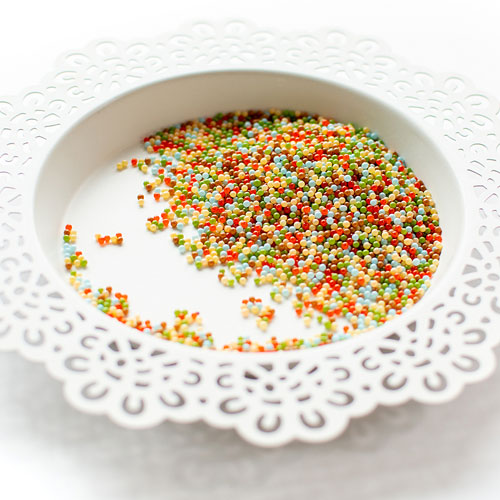 Multicolor craft beads scattered in a white ceramic bowl with a decorative white lace doily underneath, on a white background. Pretty Pink Posh Woodland Wishes Shaker Beads
