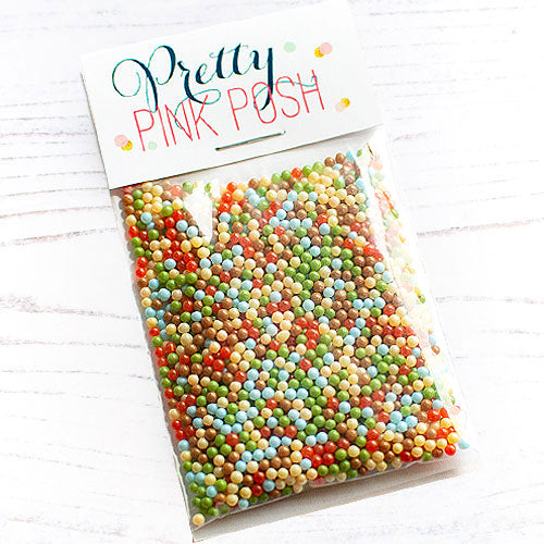 A clear plastic bag filled with many small round beads in assorted pastel and bright colors, sealed with a white header card that reads 'Pretty Pink Push.' The bag is photographed on a white wood surface. Pretty Pink Posh Woodland Wishes Shaker Beads
