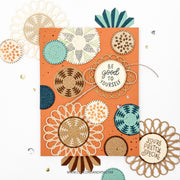 A handmade greeting card with a terracotta orange background, decorated with layered circular floral and geometric designs in beige, cream, teal, and brown. Two round wooden nameplates are attached to the card, one reading 'BE GOOD TO YOURSELF' and the other partially visible at the bottom right reading 'YOU'RE PRETTY SPECIAL'.