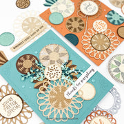 Two handmade layered greeting cards displayed on a white background, one in teal and one in orange, decorated with laser-cut circular patterns in beige, gold, brown, and teal. Small decorative tags and circular embellishments are arranged around them.