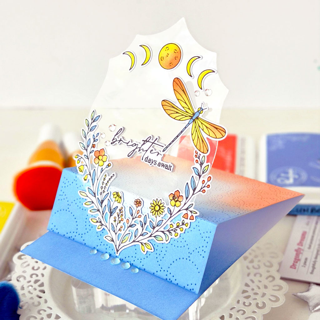 A handmade pop-up greeting card with a blue and orange gradient base, decorated with a white floral wreath, yellow flowers, a dragonfly, and moon phases, standing on a clear acrylic stand.