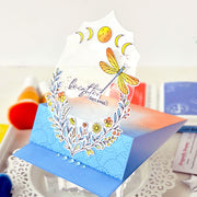 A handmade pop-up greeting card with a blue and orange gradient base, decorated with a white floral wreath, yellow flowers, a dragonfly, and moon phases, standing on a clear acrylic stand.