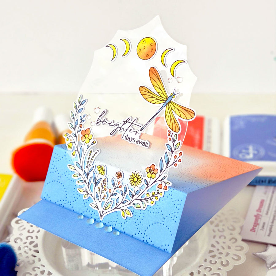 A handmade pop-up greeting card with a blue and orange gradient base, decorated with a white floral wreath, yellow flowers, a dragonfly, and moon phases, standing on a clear acrylic stand.