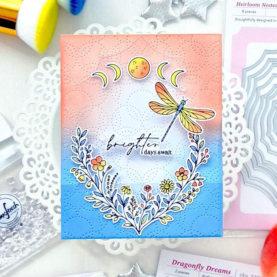 A handmade greeting card featuring a pastel gradient background that fades from peach-pink at the top to sky blue at the bottom. The card is decorated with a die-cut dragonfly, crescent moons, a sun, flowers, and the phrase 'brighter days away' in script.