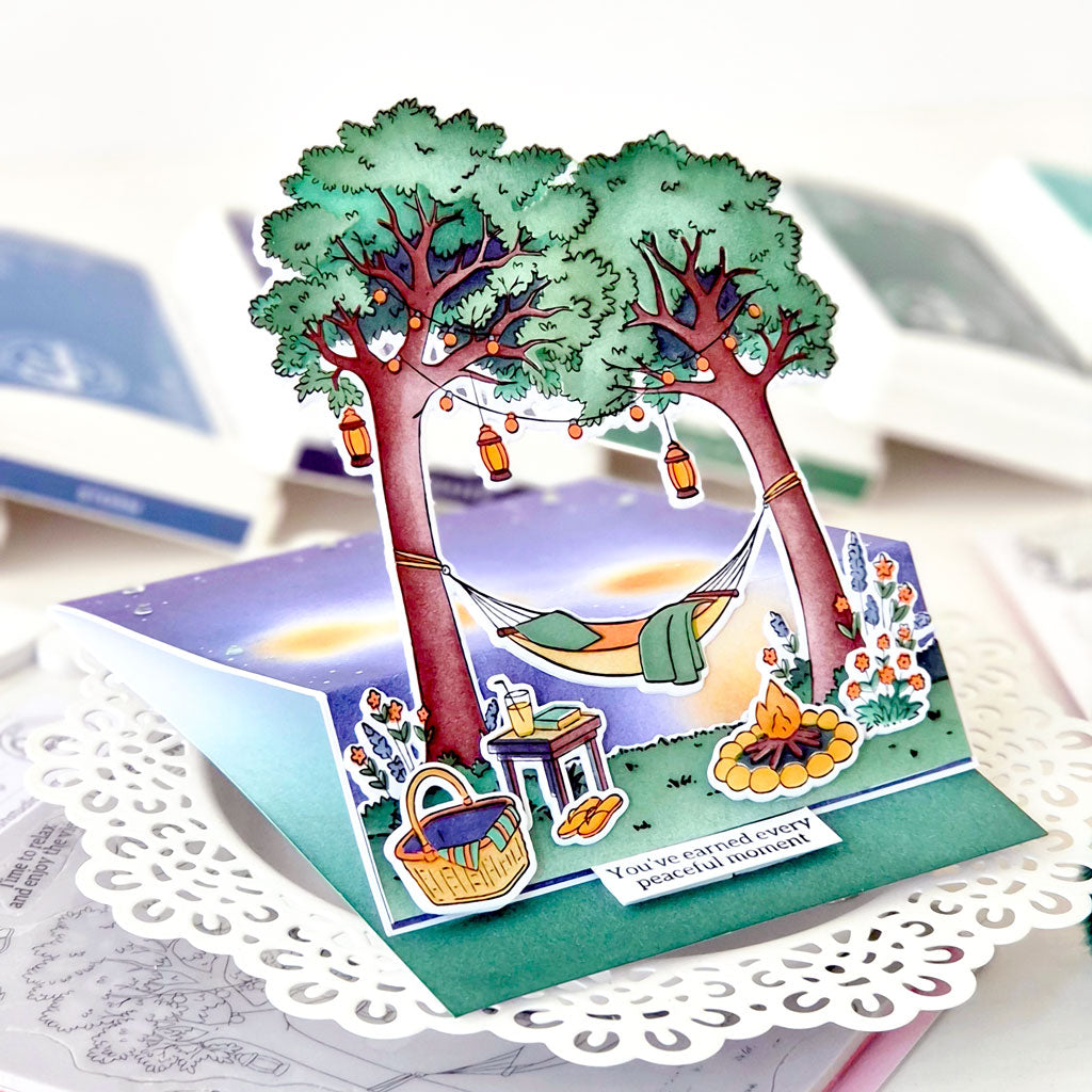 A 3D pop-up cardboard greeting card featuring two green trees, a hanging hammock, a picnic basket, and a campfire, displayed on a white doily with blurred books in the background.