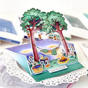 A 3D pop-up cardboard greeting card featuring two green trees, a hanging hammock, a picnic basket, and a campfire, displayed on a white doily with blurred books in the background.