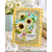 A handmade greeting card featuring three sunflower illustrations with the text 'YOU BRIGHTEN MY DAY', layered over a yellow gingham and textured beige background, styled on a lace surface with flowers and books in the background.