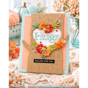 A handmade autumn-themed greeting card with a layered design featuring a kraft wood tree trunk, peach and light blue paper layers, a white heart shape with the word 'Hugs' in glittery letters, and colorful die-cut fall leaves in orange, red, and green. The card is displayed on a lace surface with decorative pumpkins and flowers in the background.