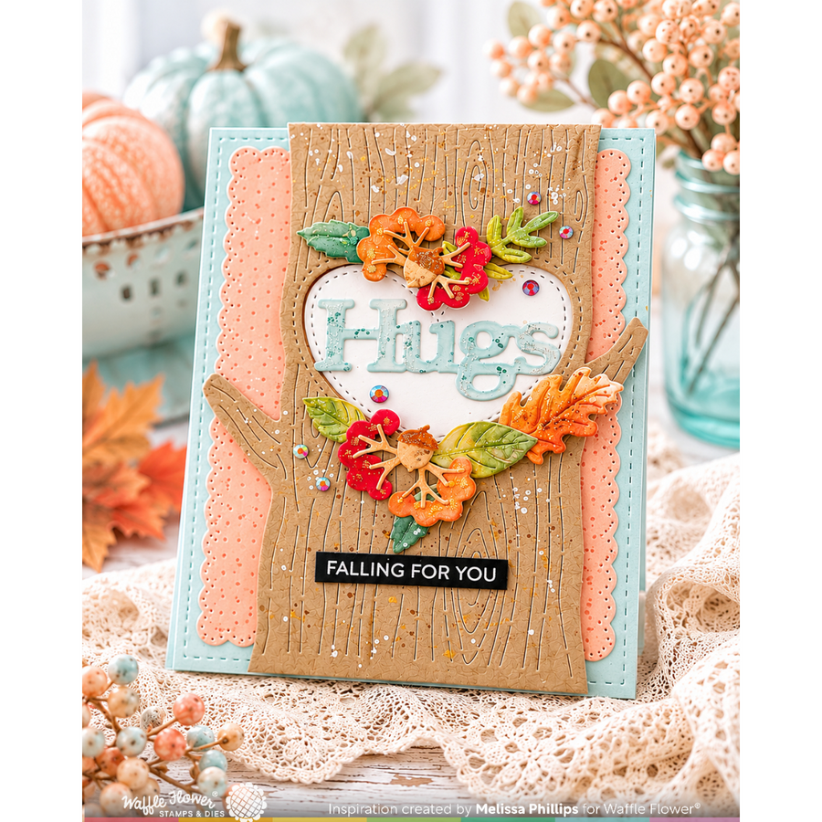A handmade autumn-themed greeting card with a layered design featuring a kraft wood tree trunk, peach and light blue paper layers, a white heart shape with the word 'Hugs' in glittery letters, and colorful die-cut fall leaves in orange, red, and green. The card is displayed on a lace surface with decorative pumpkins and flowers in the background.