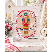 A handmade Christmas greeting card featuring a nutcracker illustration, red-and-white gingham oval border, white snowflakes, and the text 'Nutcracker Cheer' and 'MERRY CHRISTMAS'. The card is displayed on a rustic white wooden surface with holiday decorations in the background.