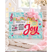 A handmade Christmas greeting card with musical notes, sheet music, and a pink ribbon bow, displayed on a rustic white wooden surface with decorative holiday props in the background.